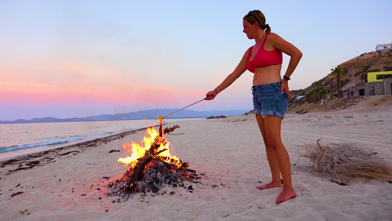 A taller burst of flame rises from the driftwood fire as the woman stands to the right, with the sunset horizon and low distant land visible across the water.