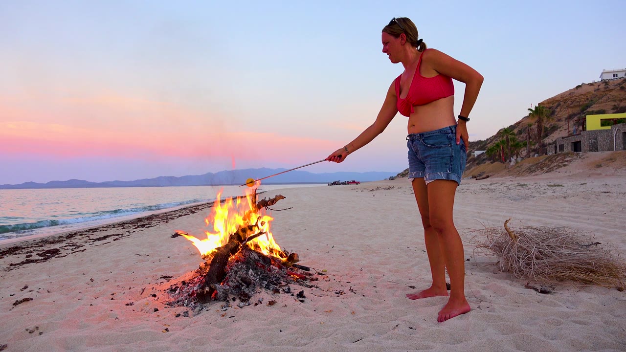 In El Sargento La Ventana, Mexico, a barefoot woman in a red top and denim shorts leans a roasting stick over a beach bonfire while the sea and pink dusk sky sit behind her.