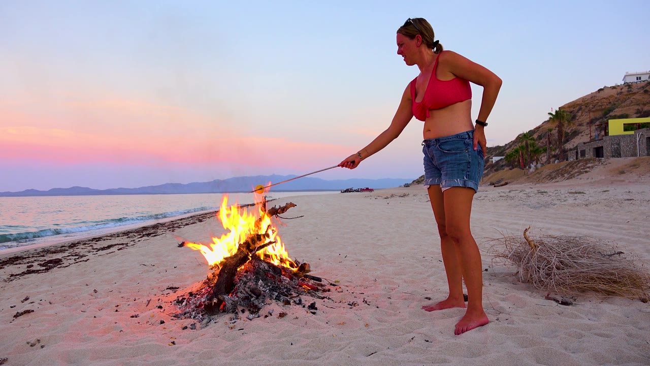 In El Sargento La Ventana, Mexico, the woman stands close to the right of the bonfire as the marshmallow extends over the flames and small waves move along the beach in the background.