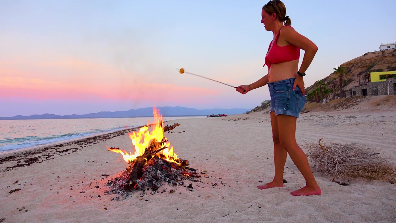In El Sargento La Ventana, Mexico, the woman has the marshmallow lifted away from the beach bonfire, with orange flames low in the center and the sunset horizon spread across the water.