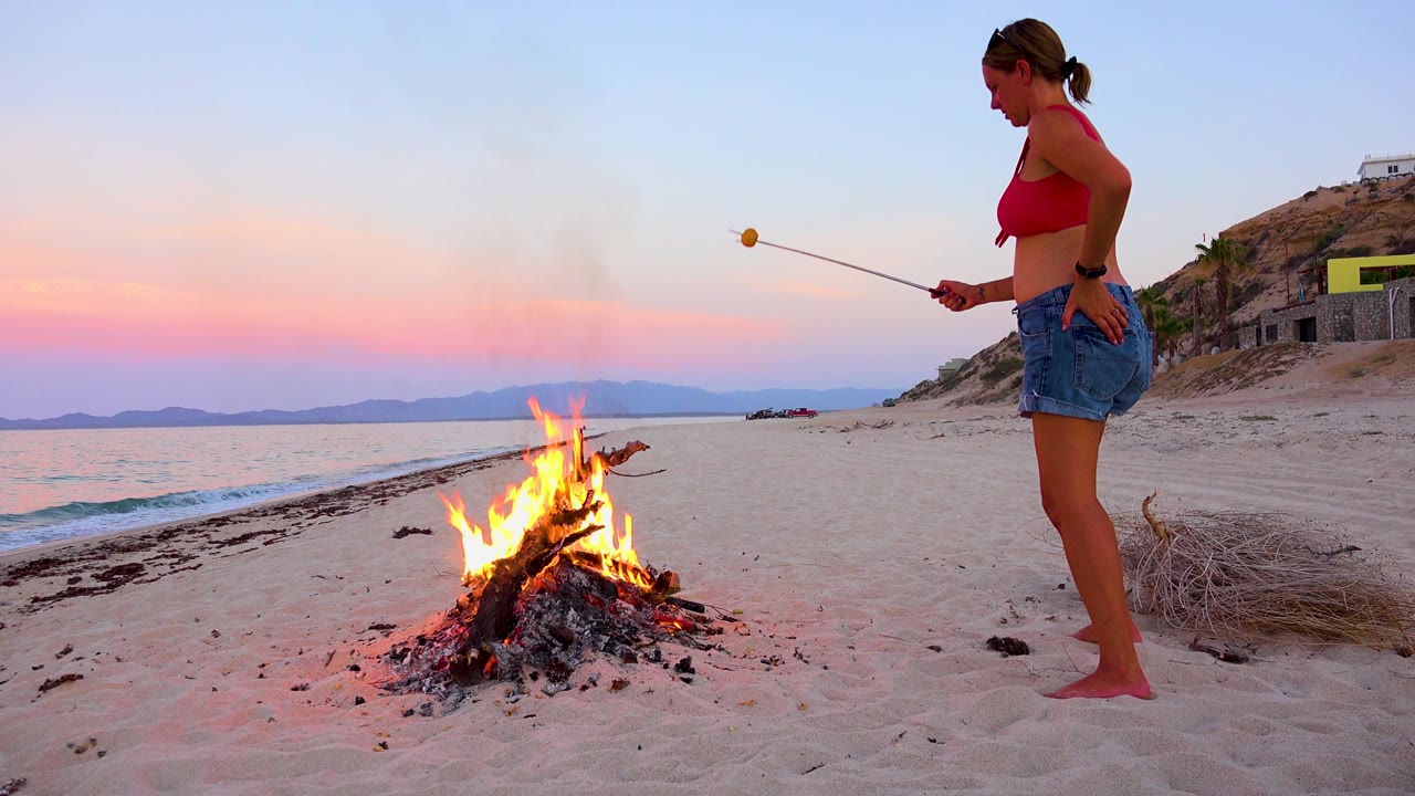 Woman watches a marshmallow over a beach bonfire at sunset.