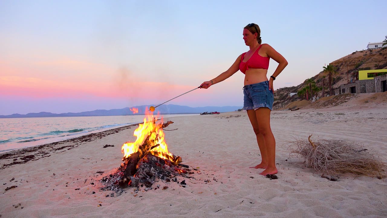 Young woman in a red top and denim skirt stands beside a bright campfire on the sand in El Sargento La Ventana, Mexico, with pink sunset light over the water.