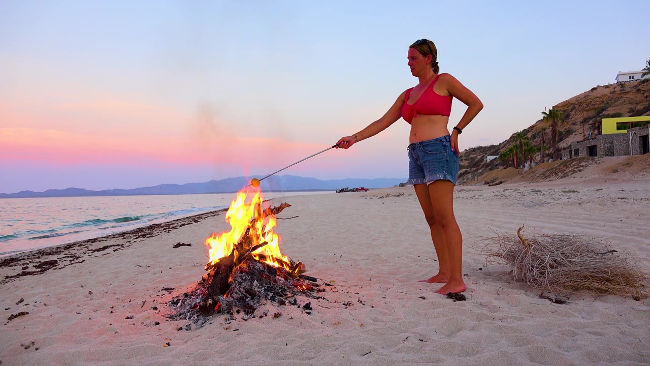 Woman holds a roasting stick over flames on a sandy beach.