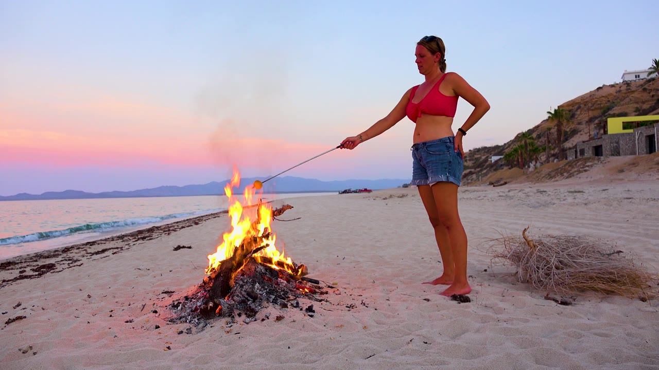 Flames flare from the beach campfire as the woman stands to the right with her arm extended, with pastel sky, water, and hillside buildings behind her in El Sargento La Ventana, Mexico.