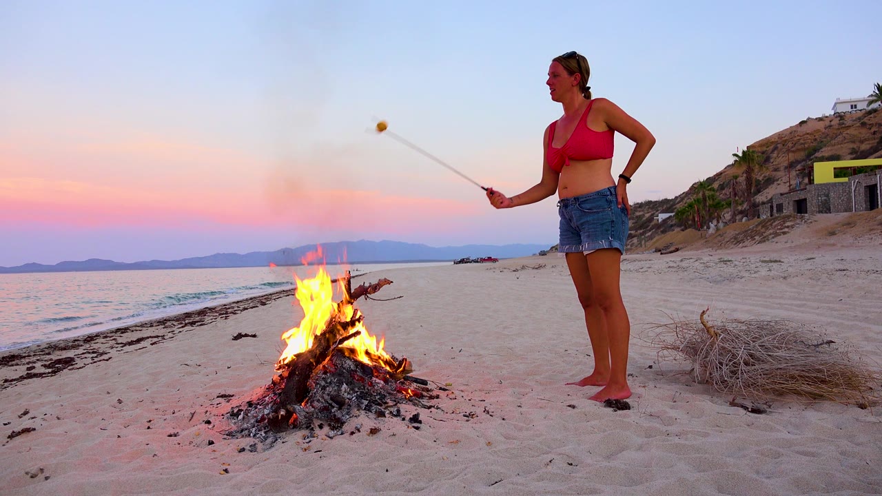At 0:00 in El Sargento La Ventana, Mexico, a woman stands barefoot on sand beside a bright bonfire with the ocean and distant hills behind her.
