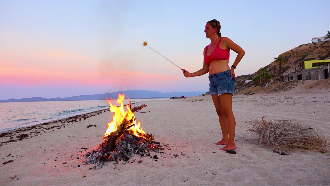 At 0:01 in El Sargento La Ventana, Mexico, the woman holds the roasting stick out over the flames while smoke rises into the pink and blue evening sky.