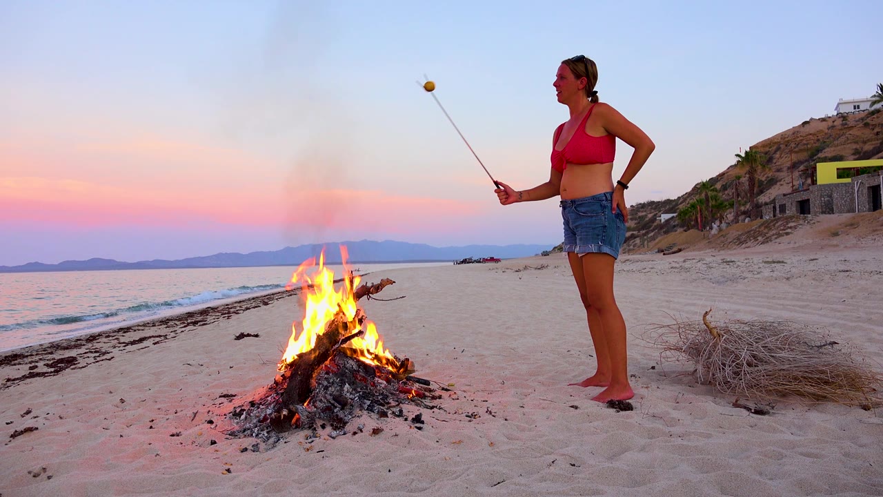 At 0:02 in El Sargento La Ventana, Mexico, the bonfire burns in the center of the beach frame while the woman stands to the right and the shoreline recedes left.