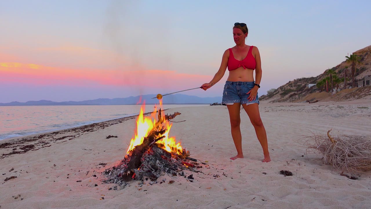 In El Sargento La Ventana, Mexico, a woman in a red top stands barefoot beside a small campfire on the sand with calm water and pink sunset color behind her.
