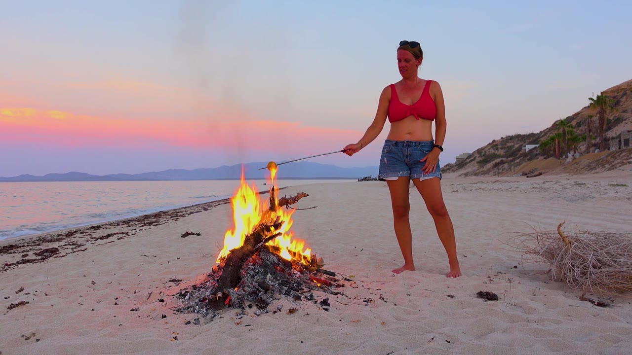 A small driftwood fire burns brightly on the beach in El Sargento La Ventana, Mexico, while a woman stands to the right holding a stick toward the flames.