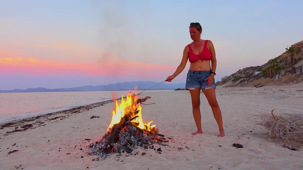 The campfire sits left of center on the sandy beach in El Sargento La Ventana, Mexico, with the woman standing farther right and a pastel horizon over the water.