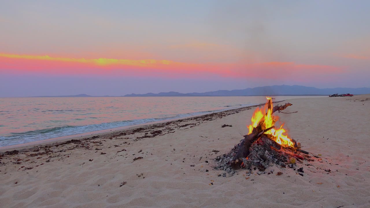 At 0:00 in El Sargento La Ventana, Mexico, a driftwood bonfire burns on sand beside gentle surf under a pink-orange horizon, with no visible people.