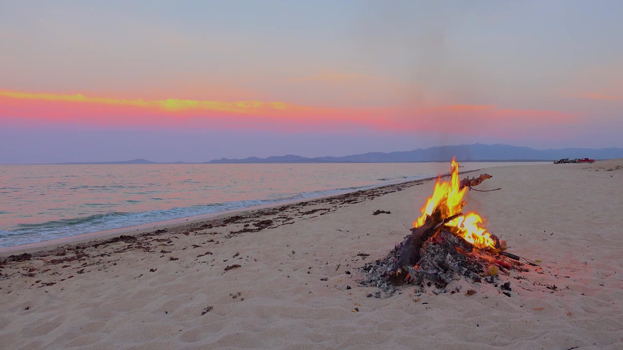 At 0:01 in El Sargento La Ventana, Mexico, flames rise from a low pile of driftwood and ash while small waves roll in behind it and no people are visible.