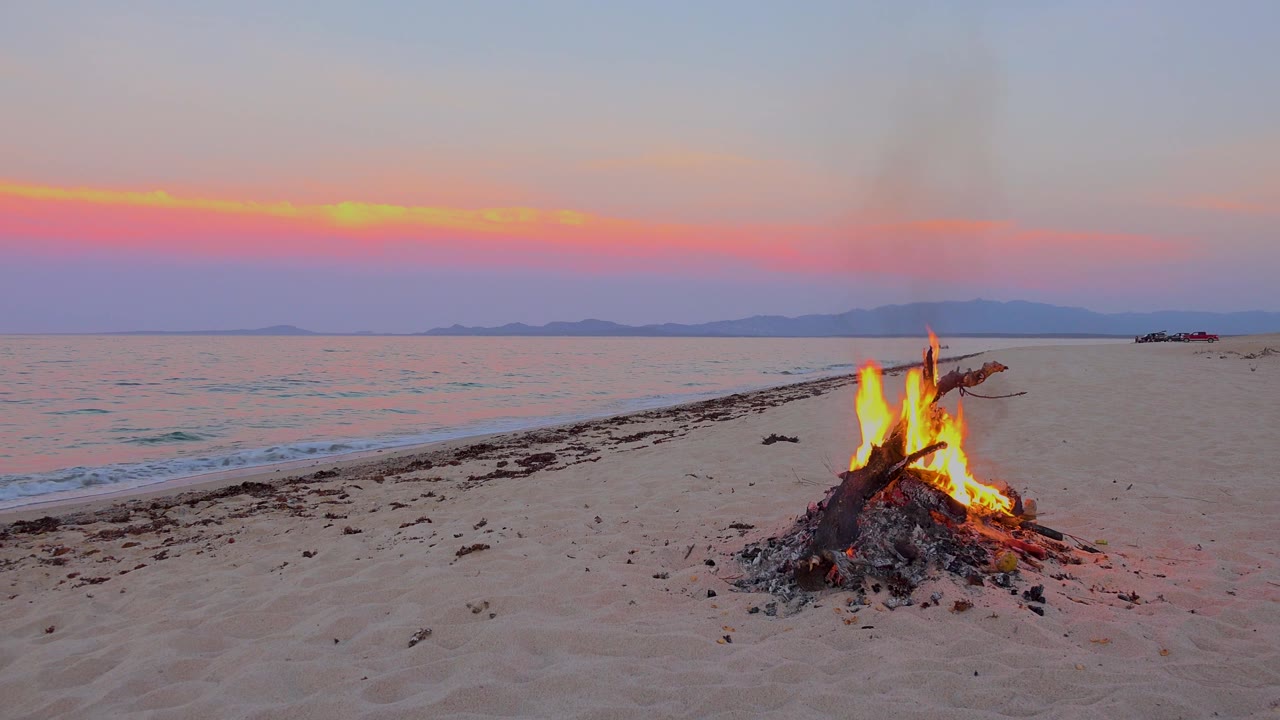 At 0:02 in El Sargento La Ventana, Mexico, the bonfire sits near the right side of the frame as a darker smoke plume lifts into the pastel sunset sky, with no visible people.