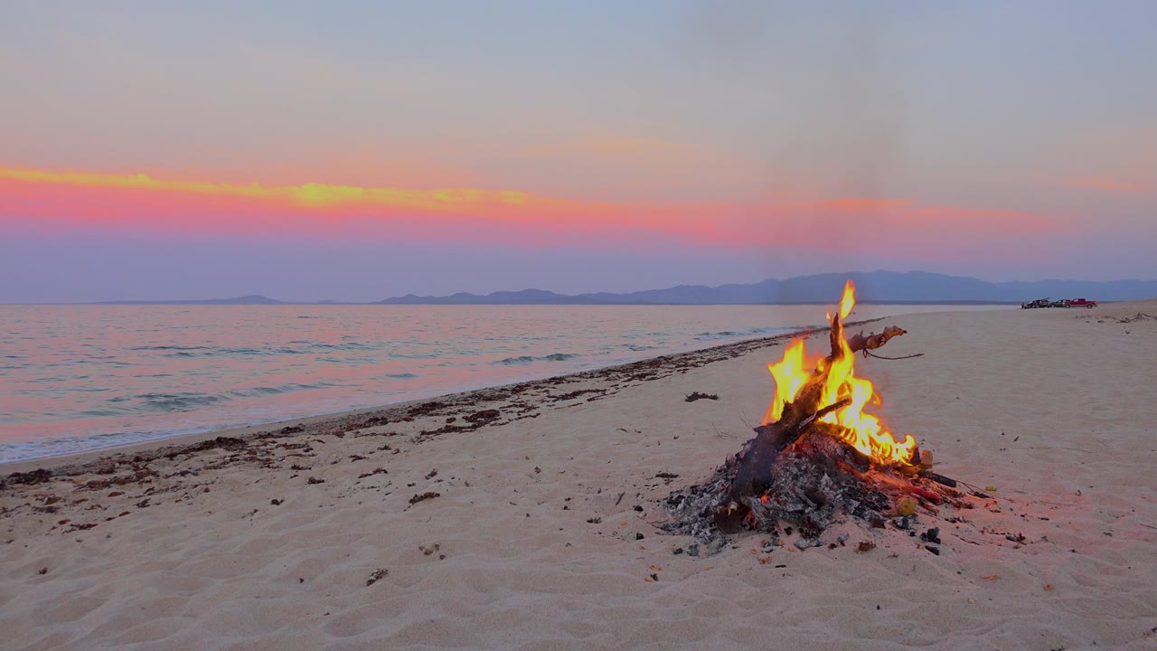 At 0:03 in El Sargento La Ventana, Mexico, the fire glows brighter in the lower right while a thin wave line reaches the sand and no visible people appear in the frame.