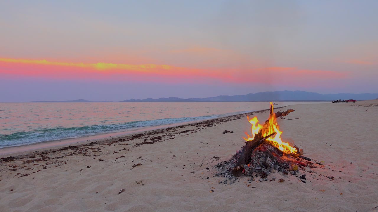 A small bonfire burns on sandy beach in El Sargento La Ventana, Mexico, with calm water, distant landforms, pink sunset light, and no visible people.
