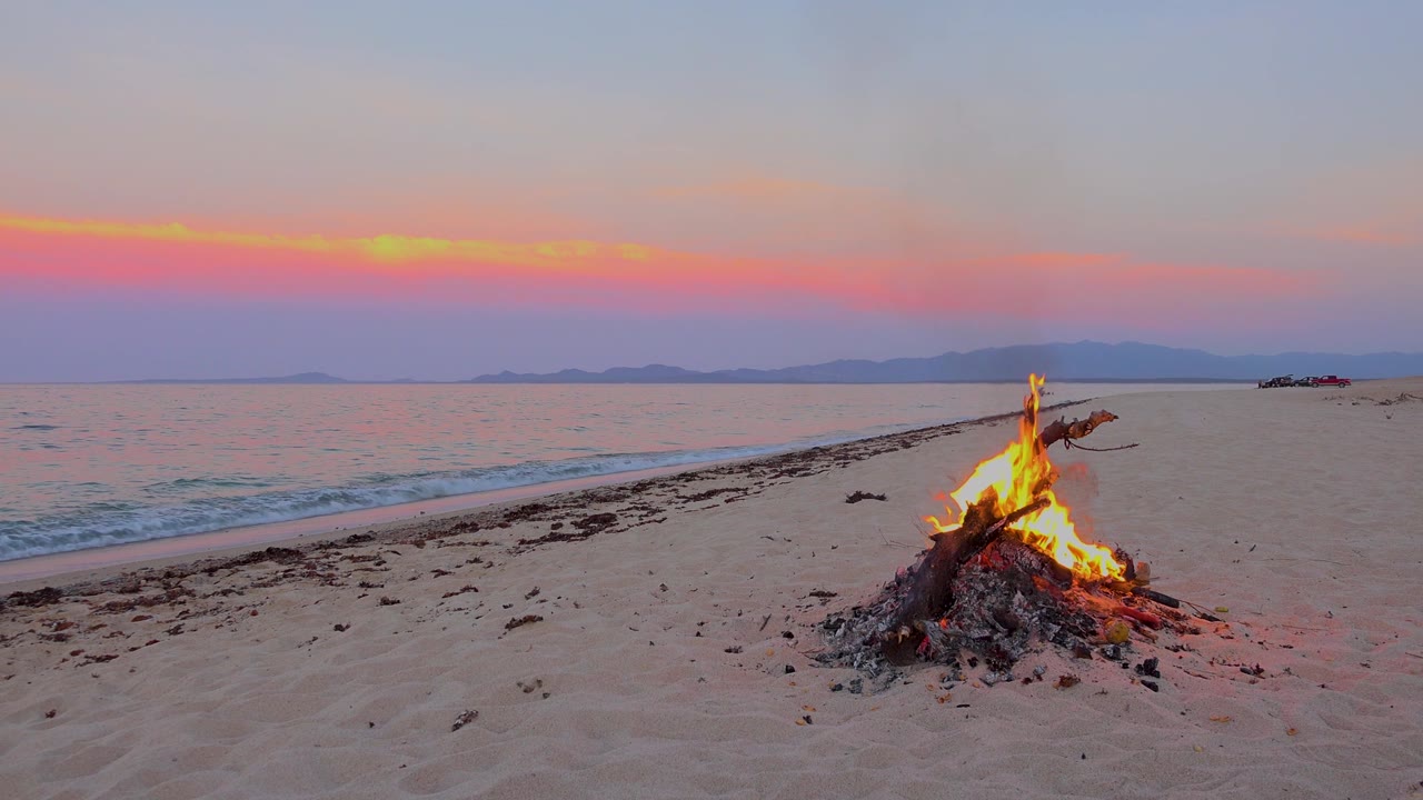 A beach fire glows on pale sand in El Sargento La Ventana, Mexico, with a low wave at left, distant hills on the horizon, and no visible people.