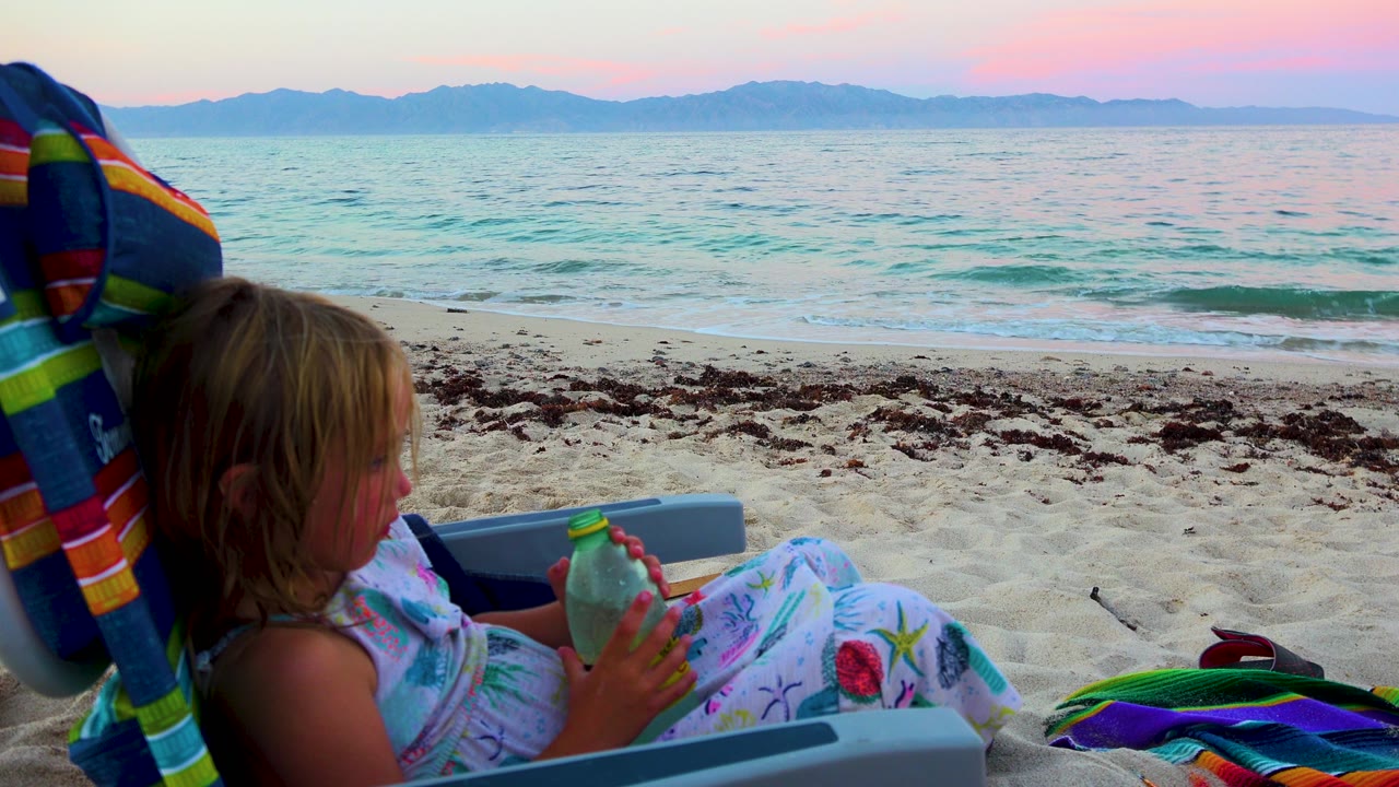 At 0:00, a young child sits in a beach chair in El Sargento La Ventana, Mexico, holding a green bottle as small waves and distant mountains sit under pink evening light.