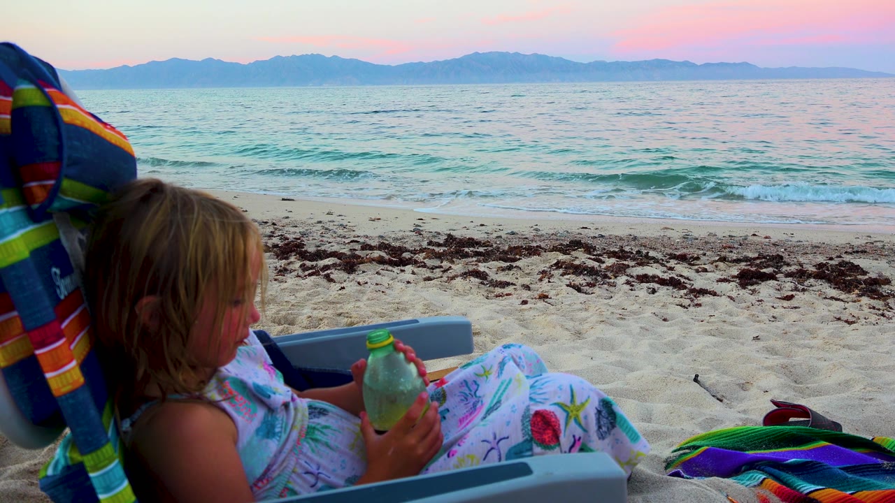 At 0:02, the child sits low in the left foreground in El Sargento La Ventana, Mexico, with the green bottle in both hands and a thin wave breaking behind a strip of seaweed.