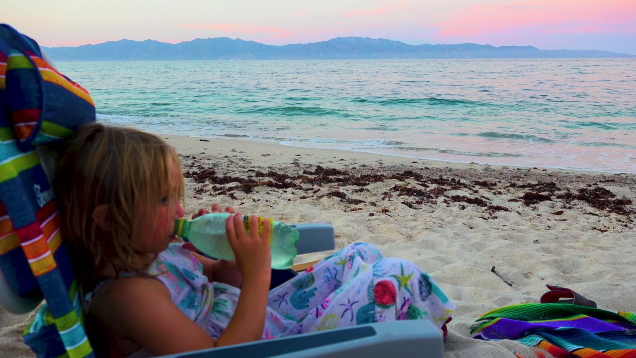 A young child in El Sargento La Ventana, Mexico drinks from a water bottle while sitting in a beach chair, with sand, surf, and distant mountains behind.
