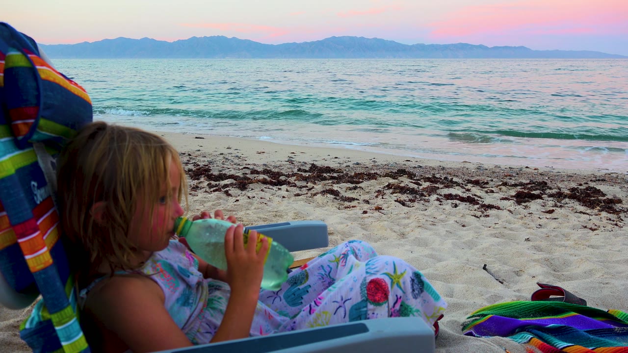 In El Sargento La Ventana, Mexico, the child is seen in profile with the bottle raised, framed against sand, shallow surf, and blue mountains under pink evening light.