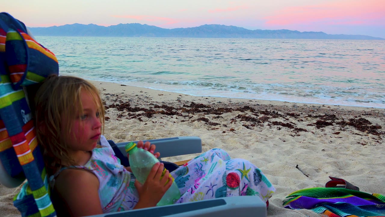 A young girl sits in a striped beach chair at El Sargento La Ventana, Mexico, holding a green bottle as small waves and distant mountains fill the sunset background.