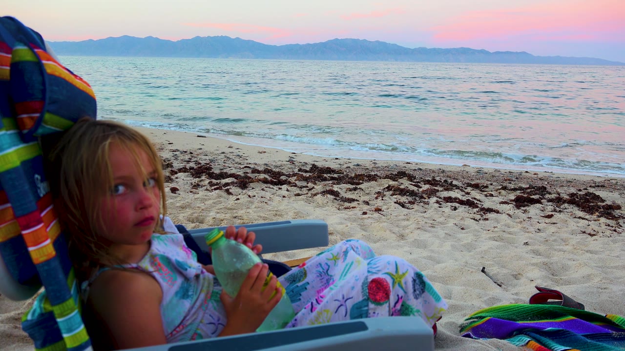 A young girl in the foreground looks toward the camera at El Sargento La Ventana, Mexico, while holding a bottle beside the beach chair and facing a calm ocean horizon.