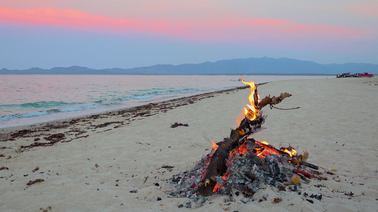 Flames rise from charred driftwood on the sand in El Sargento La Ventana, Mexico, with a pale shoreline, small wave edge, blue mountains, pink clouds, and no visible people.