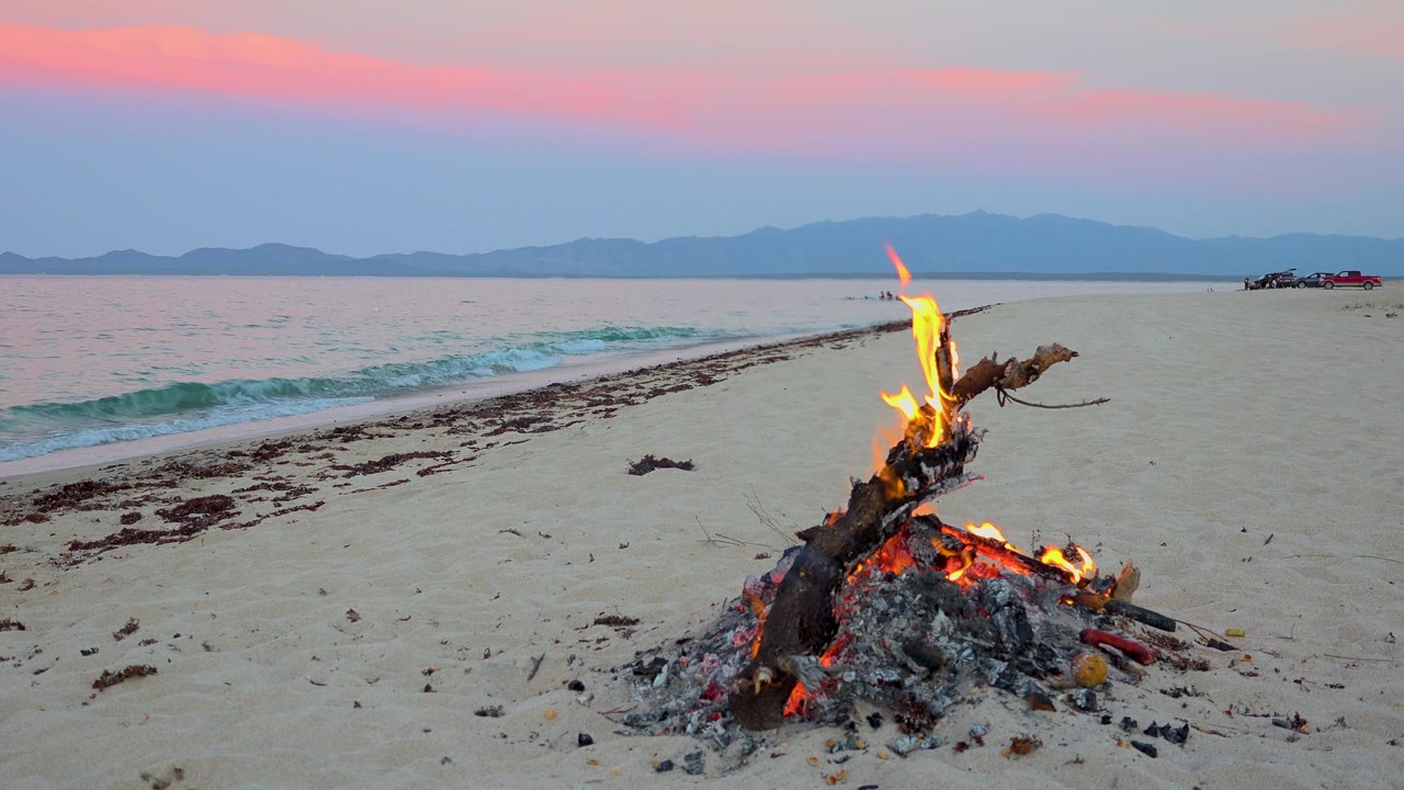 A small fire glows on the foreground sand at El Sargento La Ventana, Mexico, with the surf line sweeping in from the left, distant parked vehicles near the horizon, pink cloud bands, and no visible people.