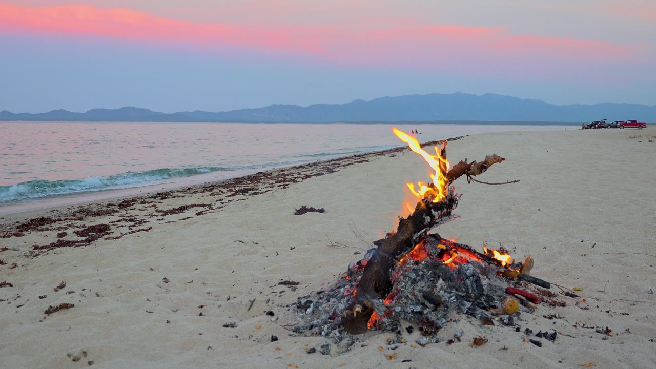 A compact fire burns low on the sand in El Sargento La Ventana, Mexico, with blue water and a pink cloud band stretching over distant mountains.