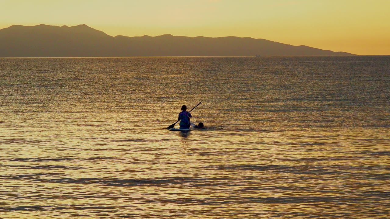 A distant paddler sits near the lower center of golden ocean water in El Sargento La Ventana, Mexico, with a dark mountain ridge and yellow sky behind them.