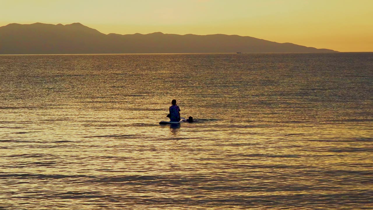 A small paddler appears farther right on reflective golden sea in El Sargento La Ventana, Mexico, with an orange-yellow sky above the dark mountain ridge.