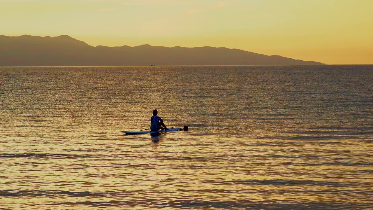 A lone person sits on a narrow boat in the water at El Sargento La Ventana, Mexico, with golden sunset light on the sea and a dark mountain coastline beyond.