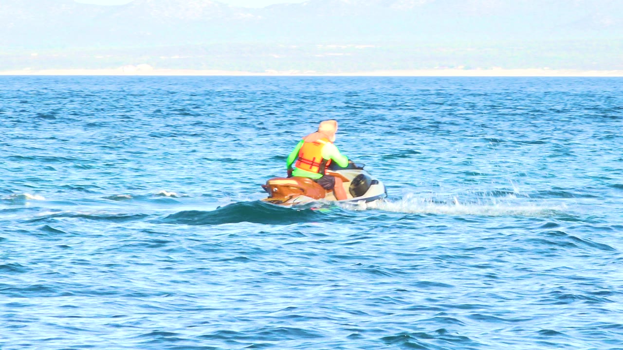 A lone rider in a bright life vest starts across choppy blue water on a jet ski in El Sargento La Ventana, Mexico, with a pale shoreline behind.