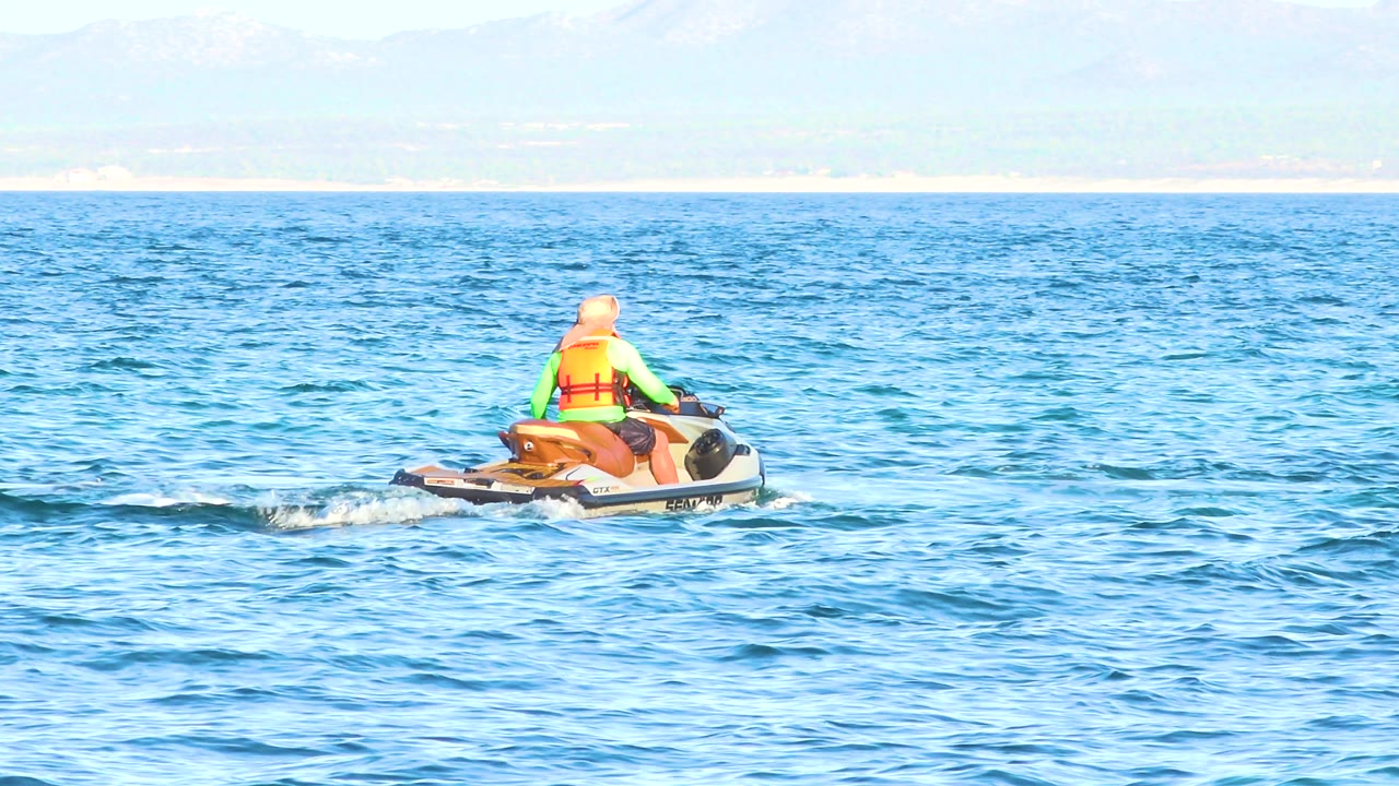 The jet ski sits farther right in frame, with the rider facing away across rippled blue water and the distant Mexican shoreline running along the horizon.