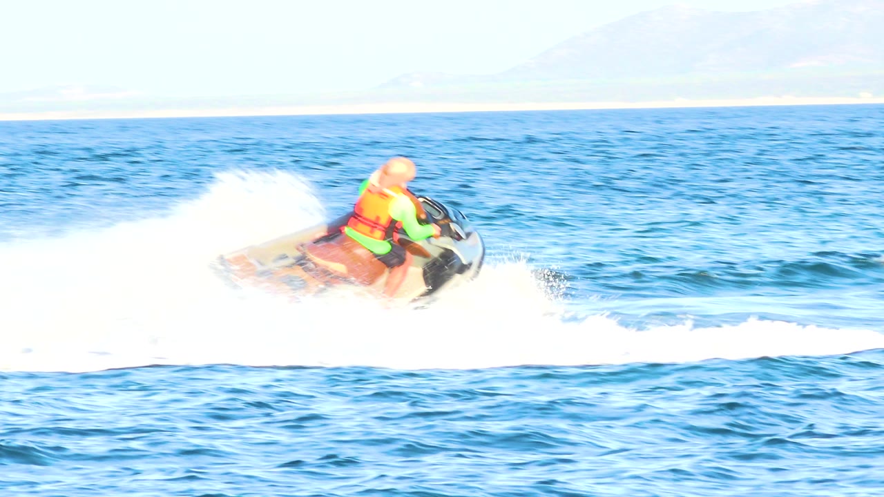 A jet ski rider moves across blue water in El Sargento La Ventana, Mexico, with bright white spray behind the craft and distant shoreline in the background.