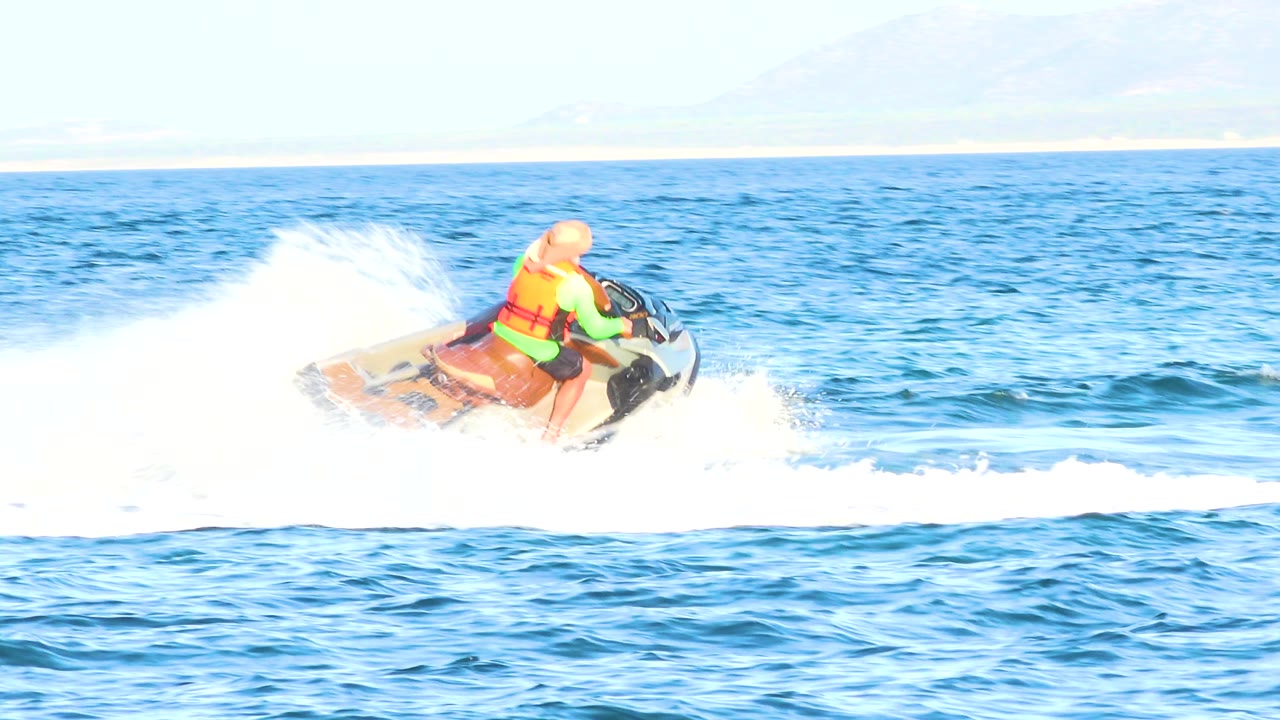 The rider on the jet ski stays midframe on the blue ocean, with a broad white spray plume and hazy landforms beyond the horizon in El Sargento La Ventana, Mexico.