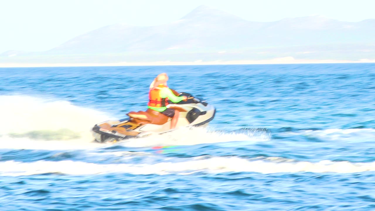 A jet ski rider in bright safety gear rides to the right across sunlit ocean water, leaving a low white wake with distant pale shoreline behind in El Sargento La Ventana, Mexico.