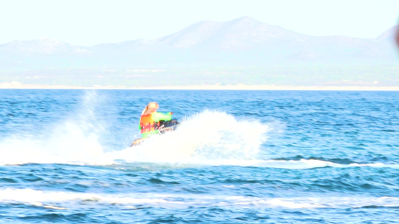 The jet ski rider is partly hidden by bright ocean spray in El Sargento La Ventana, Mexico, with blue waves in front and a pale mountain backdrop above the horizon.
