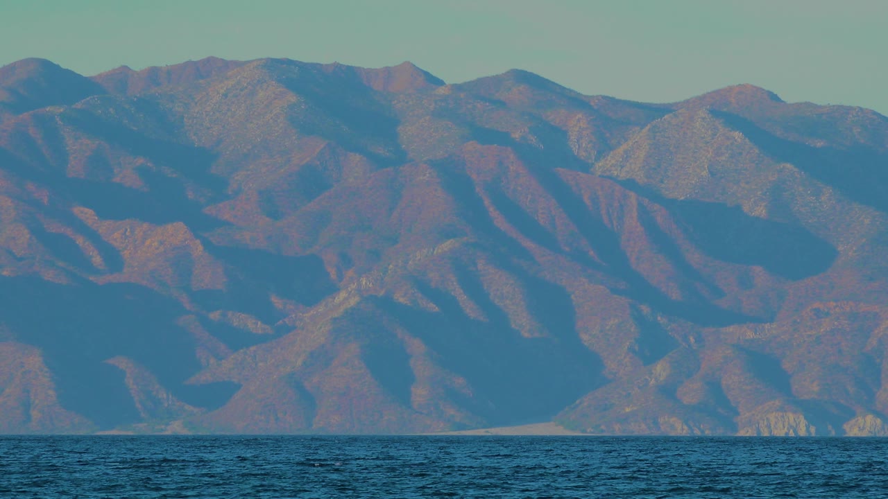 Opening frame of rugged mountains across dark ocean water in El Sargento La Ventana, Mexico, with soft sun on the slopes and no visible people.