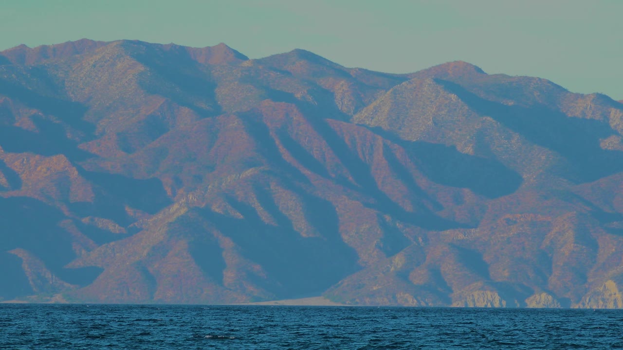 El Sargento La Ventana hillside view at 0:01, showing a dark band of water below layered tan and blue-shadowed ridges, with no visible people.