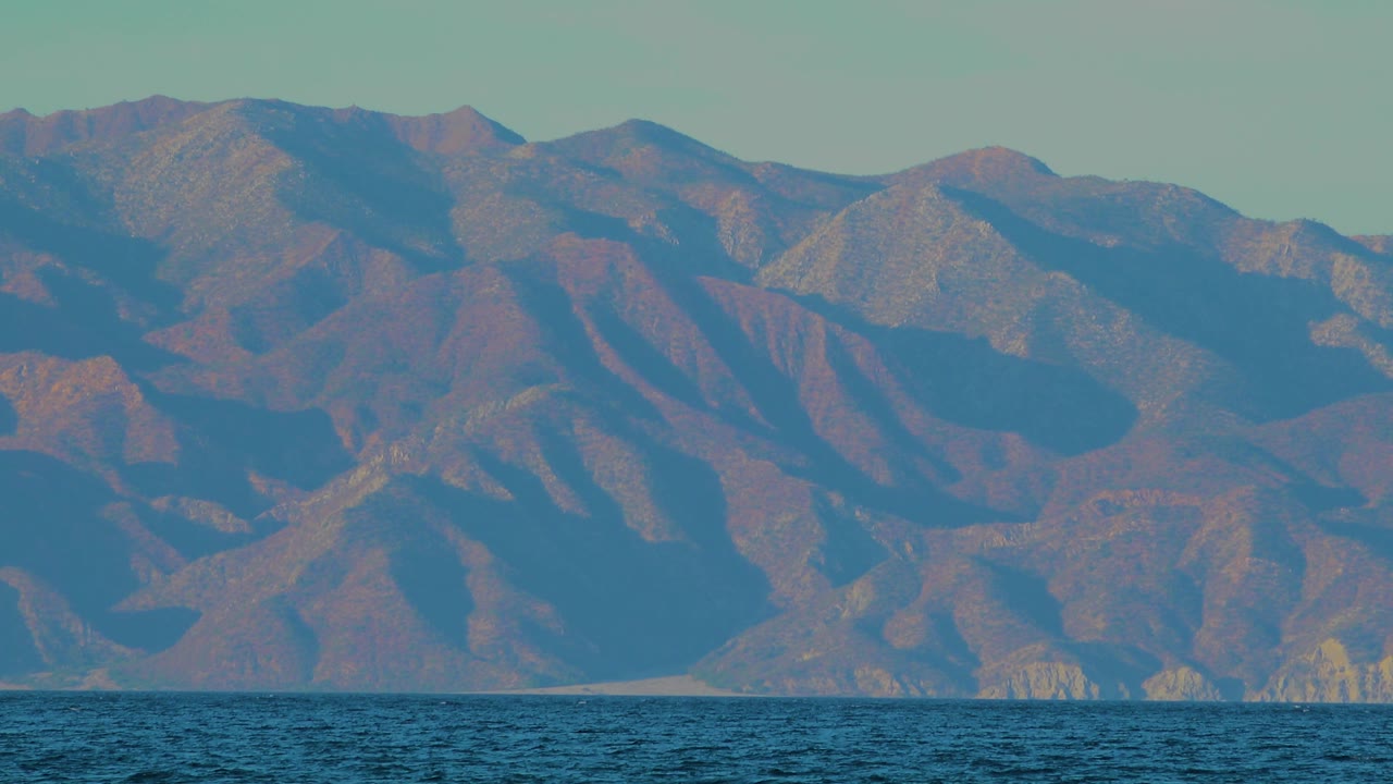 Distant rocky hills in El Sargento La Ventana rise above a dark blue waterline, with muted sun on the slopes and no visible people.