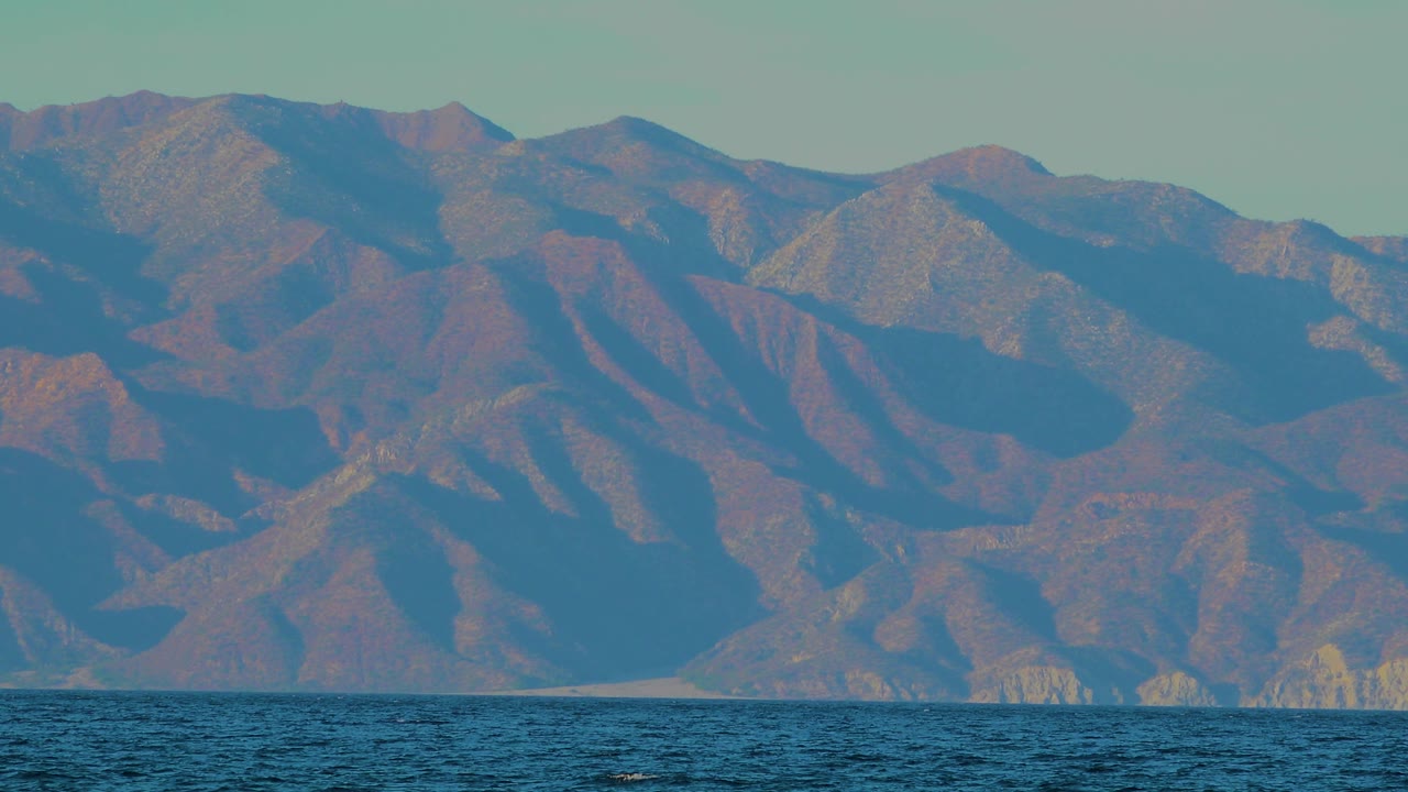 A tighter view of the El Sargento La Ventana hills shows blue shadowed slopes, tan ridgelines, a narrow strip of water at the bottom, and no visible people.
