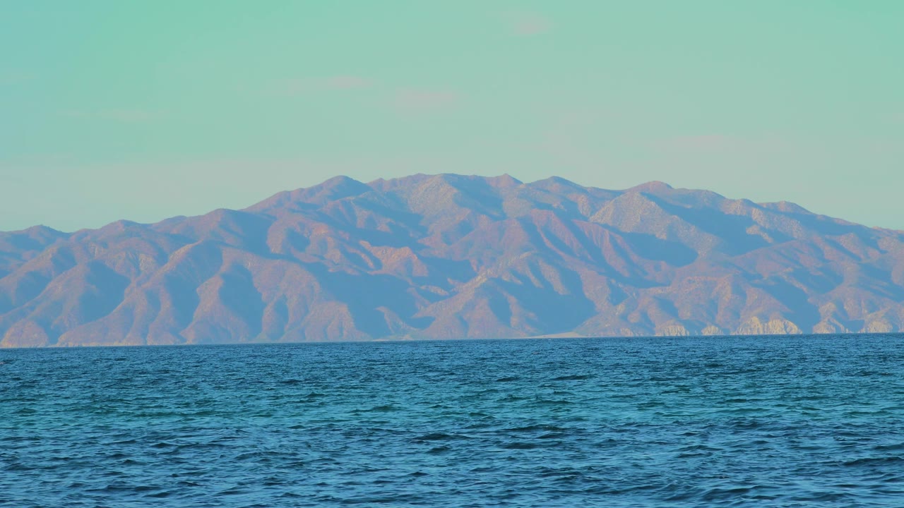 At 0:01, the ocean surface appears slightly darker in the foreground while the dry mountain ridges remain broad across El Sargento La Ventana, Mexico, with no visible people.