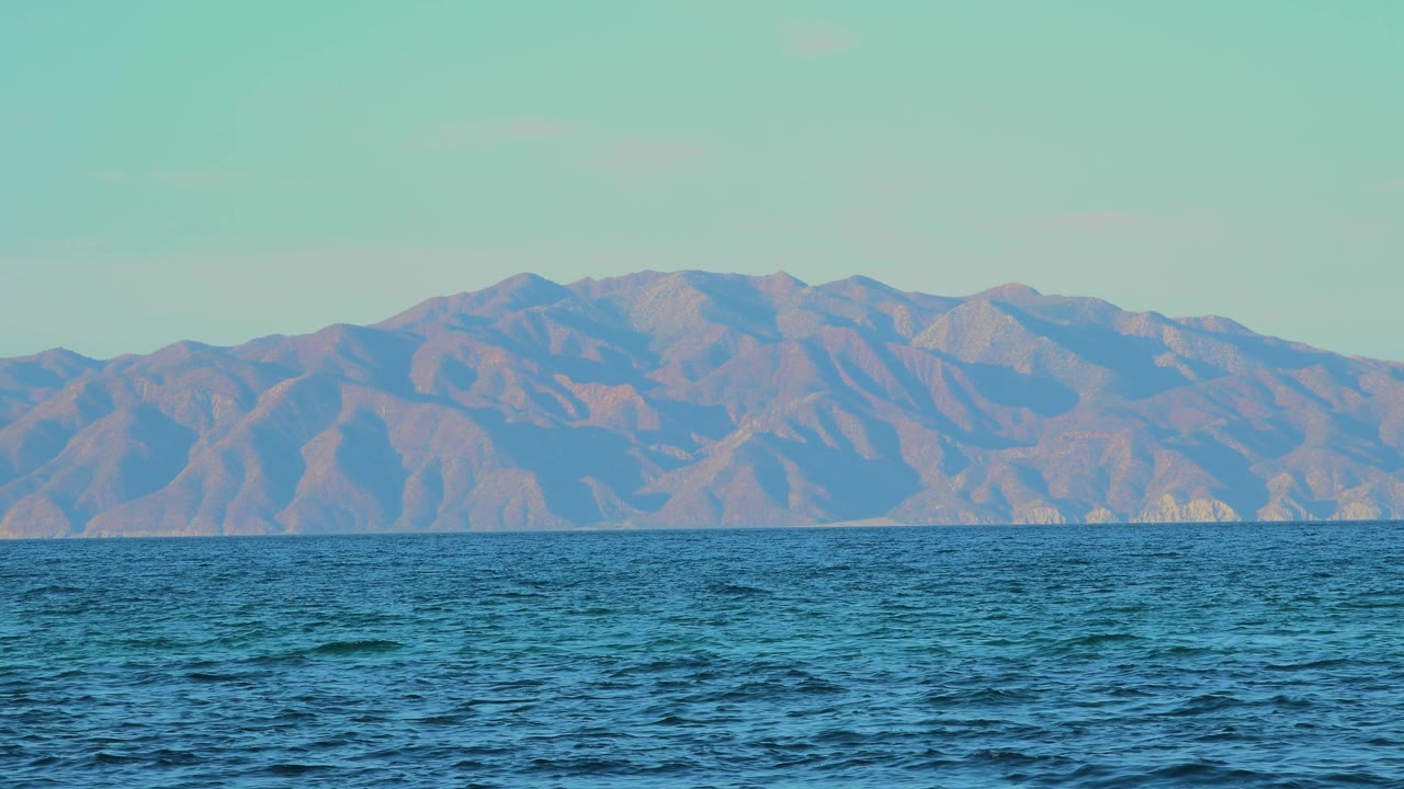At 0:02, small wind-driven waves cross the foreground while the mountain range stays centered on the horizon in El Sargento La Ventana, Mexico, with no visible people.
