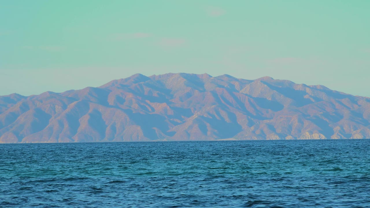 At 0:03, a wide band of blue sea runs below a long rugged ridge in El Sargento La Ventana, Mexico, with pale sky above and no visible people.