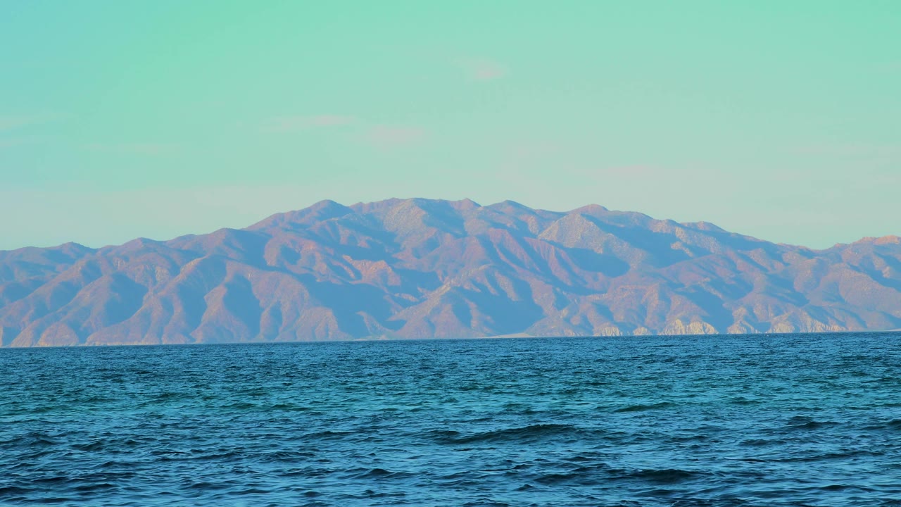 At 0:02, choppy blue water occupies the lower frame, with muted brown and blue mountains across the horizon in El Sargento La Ventana, Mexico, and no visible people.