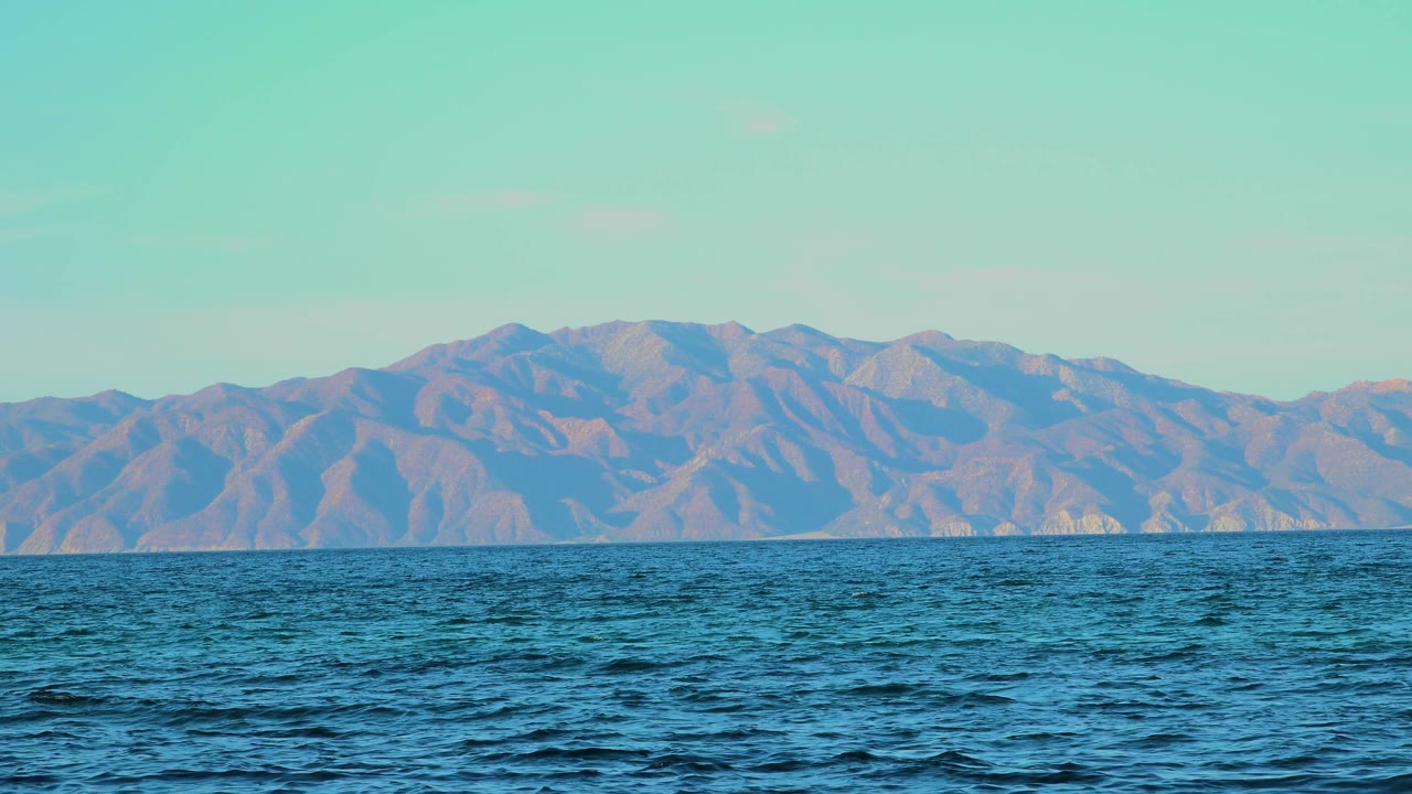Mid-clip frame with a broad band of blue water, low wave crests in the foreground, and hazy mountains stretching across the El Sargento La Ventana horizon, with no visible people.