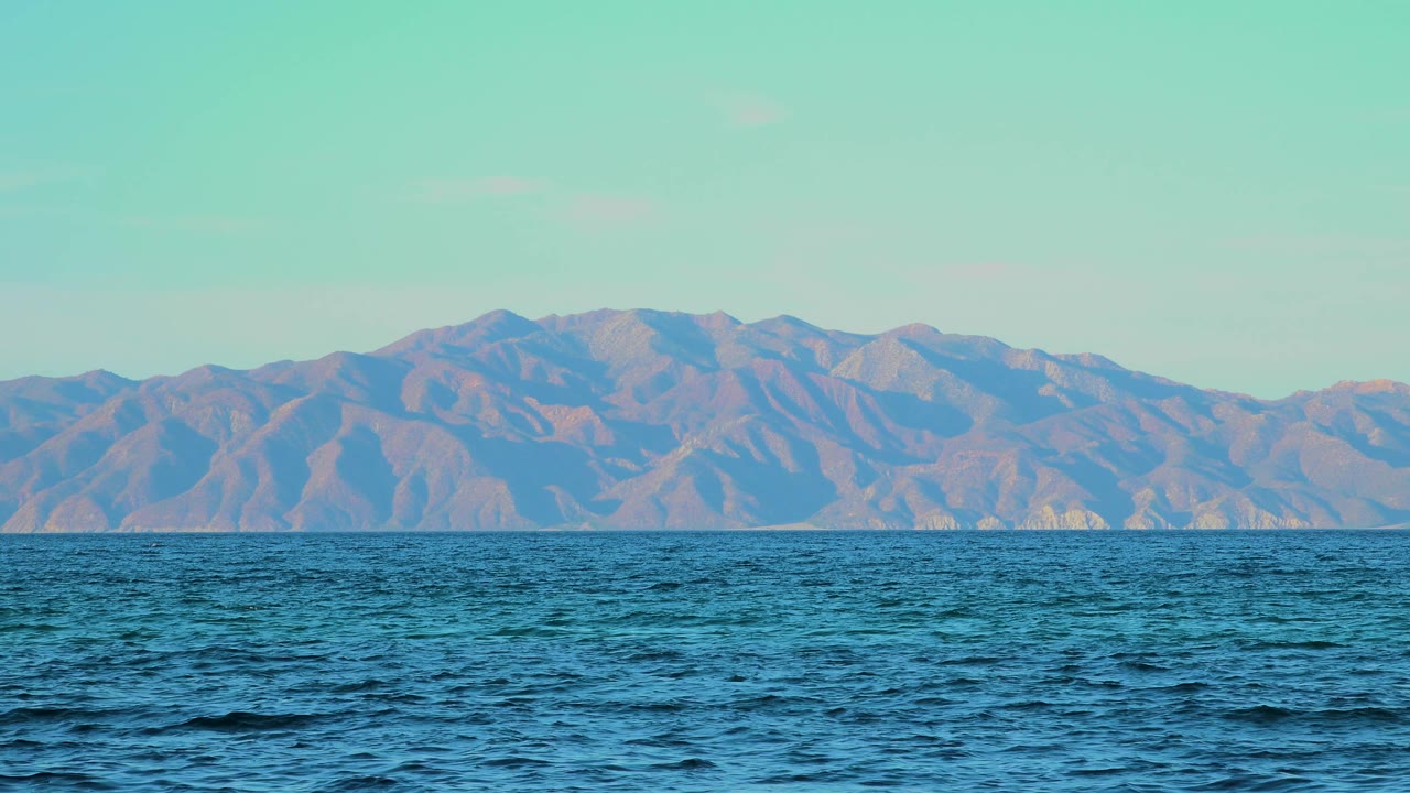 One second in, small dark ripples cross the blue water while the mountain range remains clear beneath the pale sky, with no visible people.