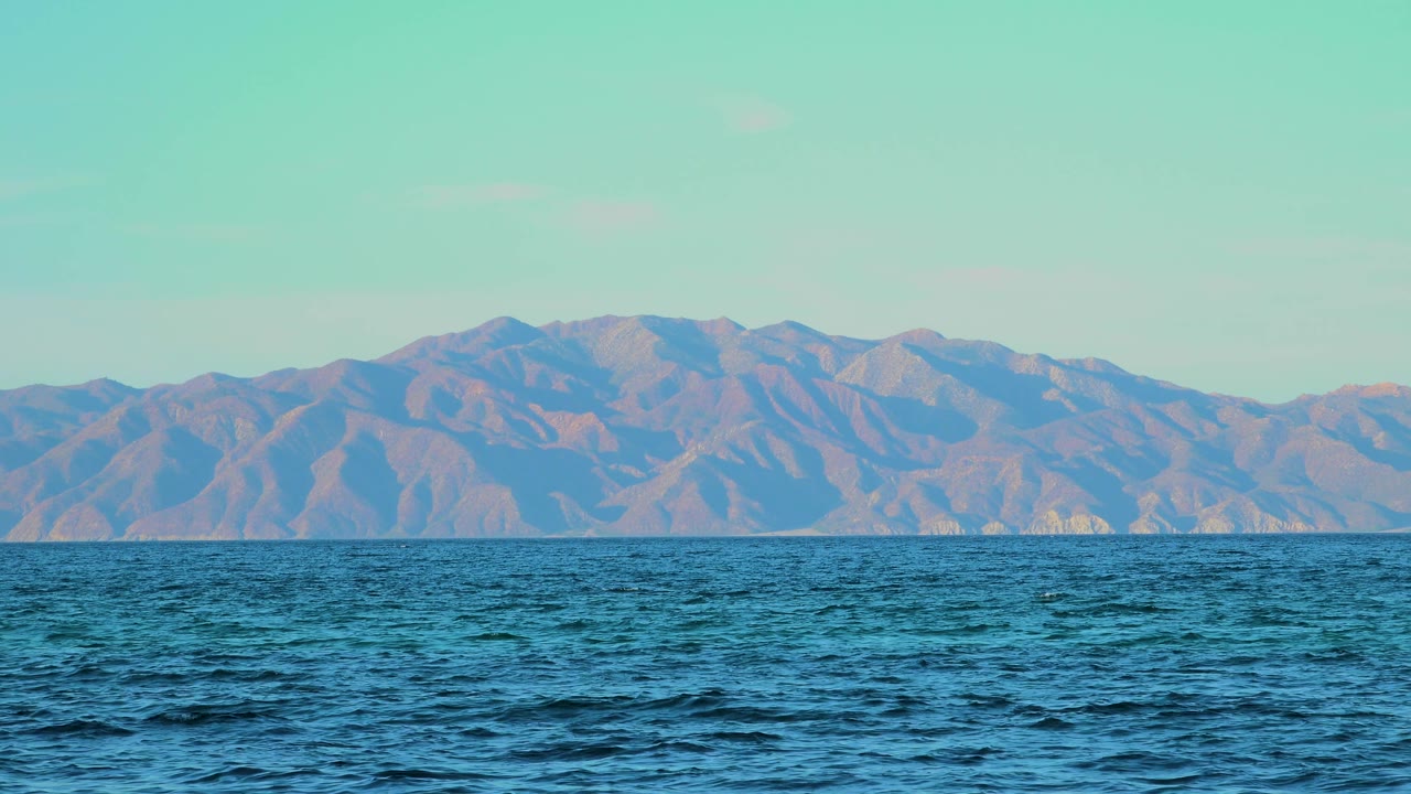 At 0:02, the frame holds a wide ocean horizon with deep blue foreground water and layered mountains across the background, with no visible people.