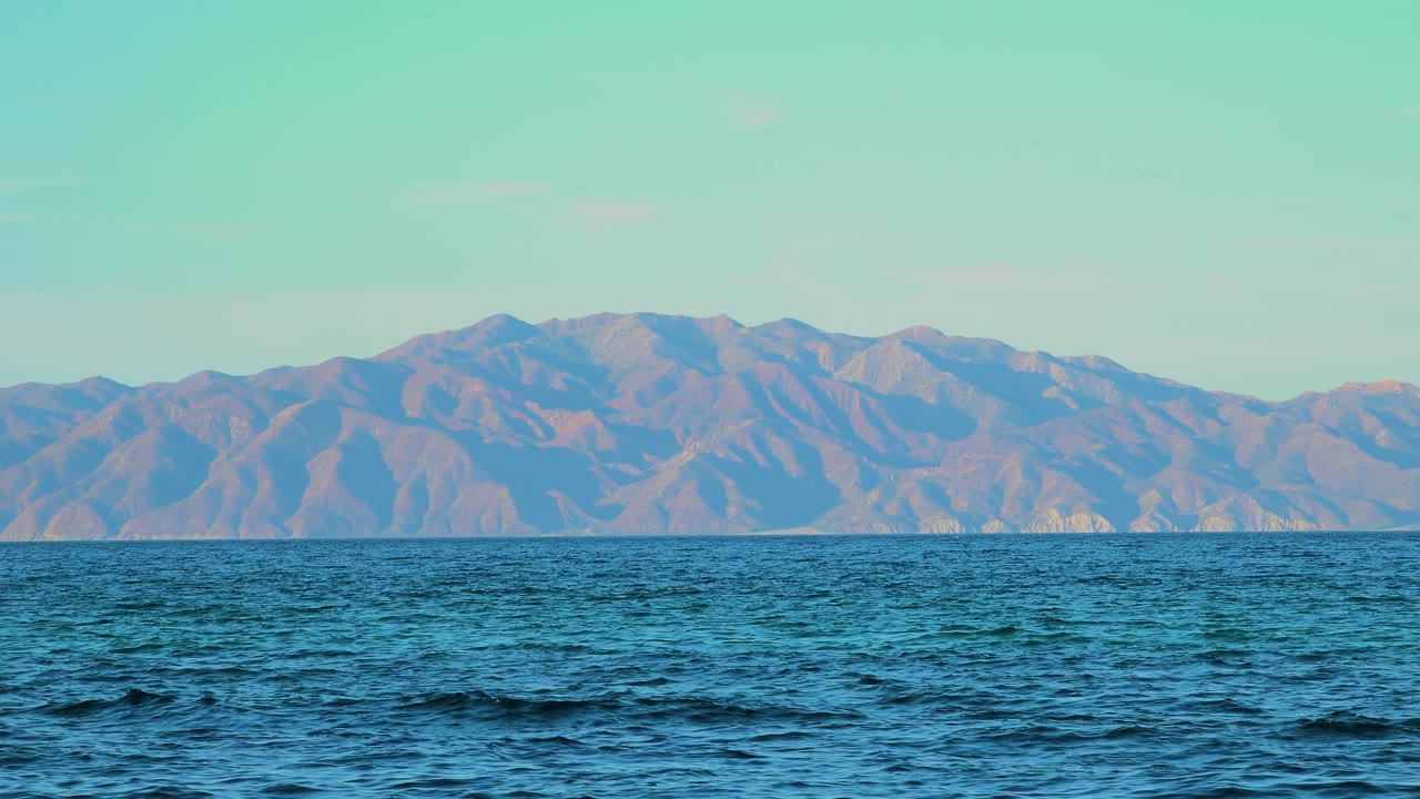 At 0:03, low waves break the ocean surface in the foreground while the distant El Sargento La Ventana mountains sit under clear pale sky, with no visible people.